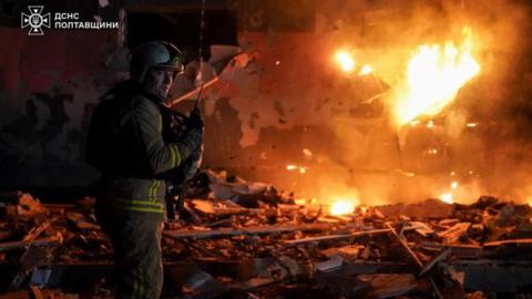 A firefighter works at the site of a building damaged during overnight Russian drone and missile strikes with flames seen in the background
