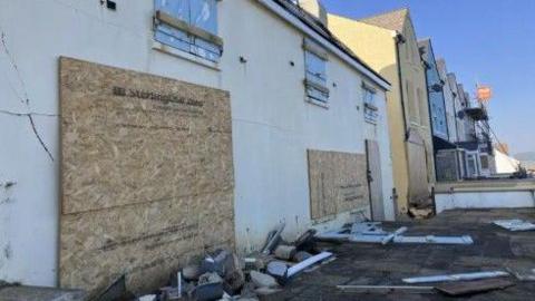 Boarded up buildings in Torcross. They were damaged by storms earlier in 2026. Rubble is everywhere on the floor.
