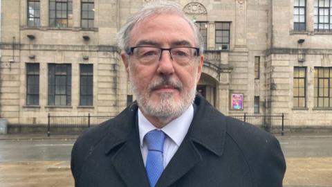 A man with white hair and glasses and a grey beard, wearing a black jacket and a  blue tie over a white shirt