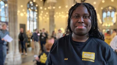 A woman stands smiling at the camera. Her top has an I WILL logo. there are people behind in soft focus and lights