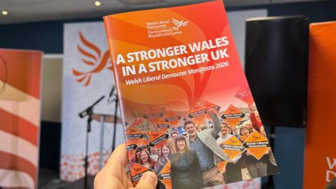 The Welsh Liberal Democrat Senedd election manifesto being held in someone's hand. The main title on the orange document is: "A stronger Wales in a stronger UK". The image on the document includes party leader Jane Dodds surrounded by party activists holding Welsh Liberal Democrat banners.