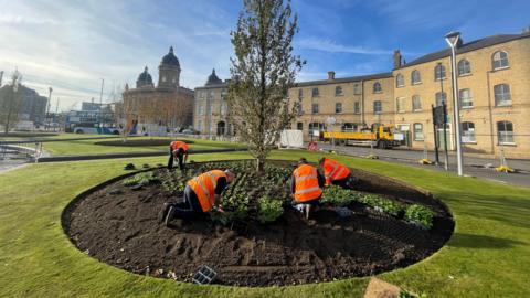 A photo of four people in orange high vis vests planting plants in a circular flower bed that surrounds a tree. Three of the people are kneeling and one is bent over. Grass surrounds the soil. There are buildings in the background. The sky is blue.