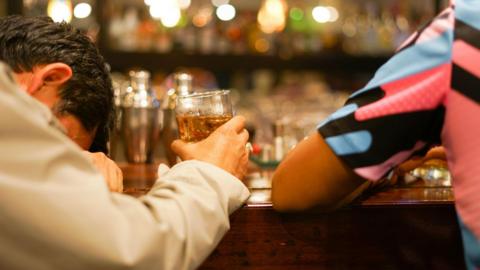 A man drunk with his head on a bar and a drink in his hand. Another man in a pink and blue shirt is next to him.