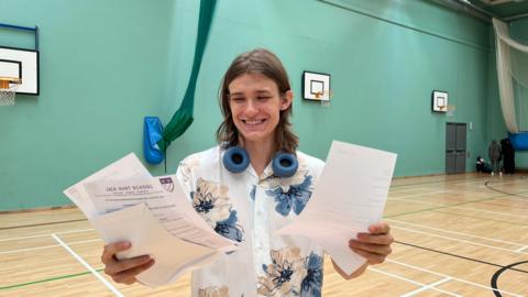 Anton is standing in a school sports hall. He is wearing a white shirt with blue and cream flowers. He has a blue set of headphones around his neck and is holding his results in both hands. He is smiling broadly.