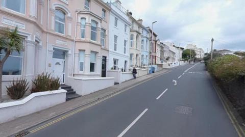 Tall colourful buildings line a road on a hill. There is a green bush on the other side of the tarmac.