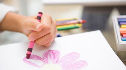 A child's hand holding a pink crayon. The child is carefully drawing and colouring in petals on a flower on a white sheet of paper. A box of crayons is just in shot. 