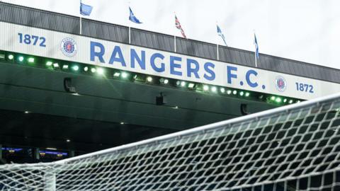 General view of Ibrox stadium under a grey sky showing the top of the goal and the main stand with flags fluttering in the wind above it