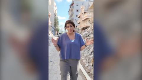 Carine stands in front of rubble as she looks to the camera while speaking
