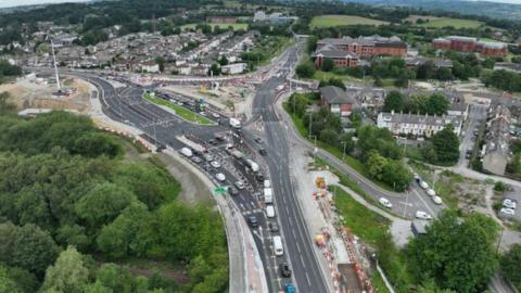 A drone shot shows the Dawson's Corner roundabout in Pudsey, Leeds. It is taken mid-construction, with cones and temporary lanes visible. Some cars are queueing at the traffic lights.