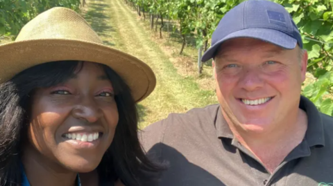 A woman in a straw sunhat and a man in a polo shirt and a baseball cap stand in a vineyard
