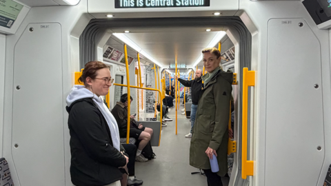 Two undercover police officers stand in between two Metro train carriages. One of them (left) is wearing a grey hoody, and grey sweatpants, and has a small black earpiece in her ear. She is smiling at a passenger who is sitting down opposite her. On the right, her colleague is wearing a long green jacket and holding a purple metal water bottle. The Metro train is in motion and inside a black tunnel.