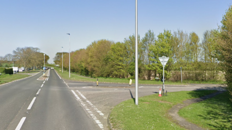 A single carriageway is on the left and a junction leading to the road is on the right. Along the roadside are several trees and silver lampposts.