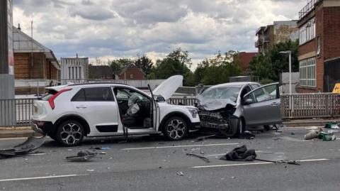 A white car and a grey car are badly damaged from the impact with debris scattered across the road.