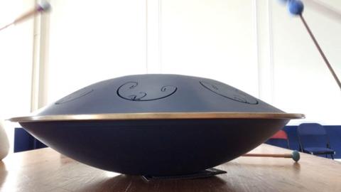 A Rav Drum on a wooden table. There are come chairs in the background.
which makes a therapeutic sound – work wonders in calming children who 