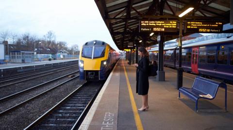 A woman in a dark coat and high heels is standing on a train station platform looking at her phone as a train is coming in.  Above her are electronic boards displaying train times.