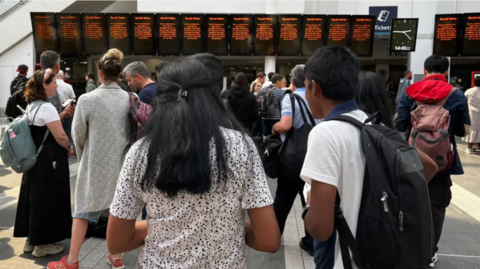 The backview of a mixed group of people who are all standing in Birmngham New Street station looking at a display board