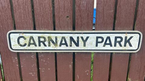 A close up of a brown fence has a white sign on it, with black writing. The long rectangular sign has the words 'Carnany Park' in block capitals, on it.