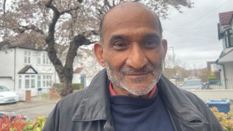 Kamal Shah, a man of South Asian heritage with a greying stubbled beard and a black jacket, standing in the front garden of a house on a residential street
