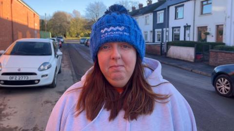 Melissa McComb - a woman stands in the middle of a street where terraced houses are behind her and looks at the camera. She has medium-length brown hair and is wearing a blue beanie had with 'Wild Atlantic Way' embroidered in white and a grey hooded jumper.