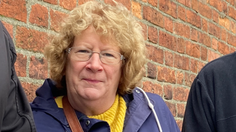 A woman with ginger curly hair, glasses, a blue coat and yellow jumper stood in front of a brick wall