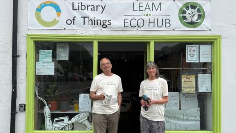 A man and woman in white t-shirts stood outside a shop with lime green window frames. The sign above the shop reads Library of Things and Leam Eco Hub