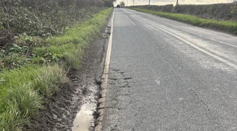 A close up of a puddle in a pot hole on the edge of a road that is cracking at various points.
