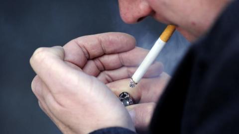 A man lighting a cigarette that is in his mouth. He has cupped his hands, where he is holding a lighter, and he is smoking a lit cigarette. You can only see his mouth and nose, and he is wearing a dark top. 