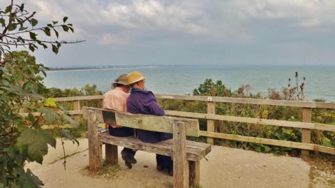 Two gentlemen sit on a bench overlooking a beach. A fence runs around the front of the bench with hedges and gorse behind. The sea can be seen in the distance. 