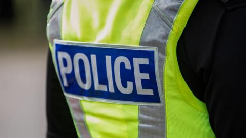 The back of a police officer, who is wearing a black jacket and a high-visibility vest, with the words police printed on.