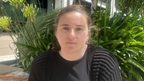 Verity O'Connell is sat in Trenton Square with flowers behind her. She is wearing a black cardigan and a black top. She has brown hair and brown eyes. 