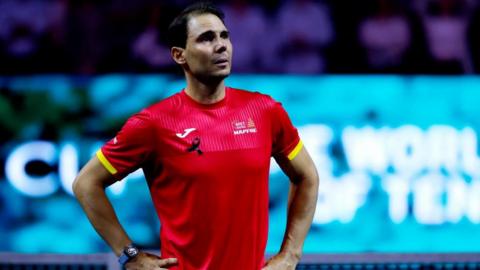 Rafael Nadal waves to the fans after loosing his singles match in the Davis Cup tie between Netherlands and Spain on 19 November 2024 in Malaga. He is wearing a red Spain top and has his hands on his hips