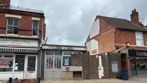 A brick terrace with a large gap in the terrace where the former bank used to be. The former bank metal and glass doors can still be seen at the front with wood boardings over the rest of the front of the building site. 