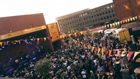 A large crowd is watching a band playing on a stage in front of the Fire Station venue in Sunderland. String of pink, yellow and red flags are hanging in the air.