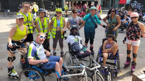 A group of eight people in roller skates standing behind three people in wheelchairs. 