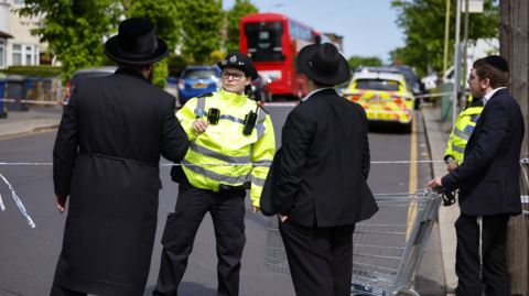 A female police officer in yellow hi-vi jacket speaks to three members of the Jewish community in traditional Orthodox Jewish clothing (black top hat; black long coat). One young man, to the right of the frame, is pushing a supermarket shopping cart