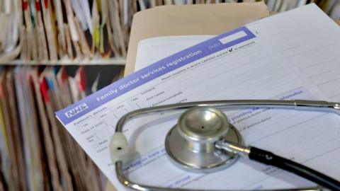 A stethoscope lies on a GP registration form. Files can be seen in the background.