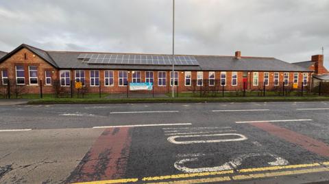The outside of a one-storey primary school with black fencing running around the outside of the building. Solar panels are attached to the roof.