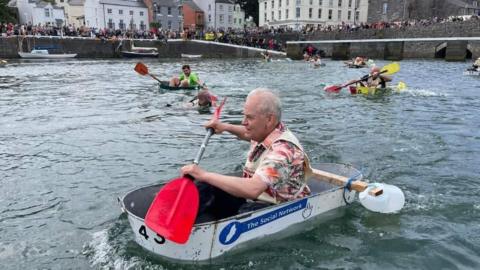 A man in a tin bath paddles in Castletown Harbour, there are other competitors in the race behind him in the water. One man's bath has sunk and he is in the water. The harbour walls are lined with hundreds of spectators.