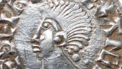 A close up of a silver penny of Ethelred II, king of England. It shows a profile portrait of the king with a mass of hair like a long Mohican. Letters from his name, struck on the circumference can be seen.