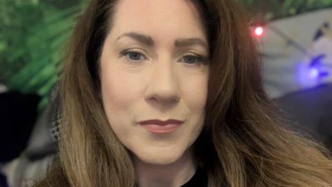 A woman with long brown hair looking at the camera. Her lips are closed and she does not smile