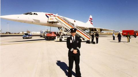 John Tye standing in front of Concorde plane Alpha Foxtrot on the runway. He is dressed in his black pilot uniform and a black cap. He has his hands crossed in front of him. Behind his, others can be seen fixing up the plane, with a bright blue sky above them.