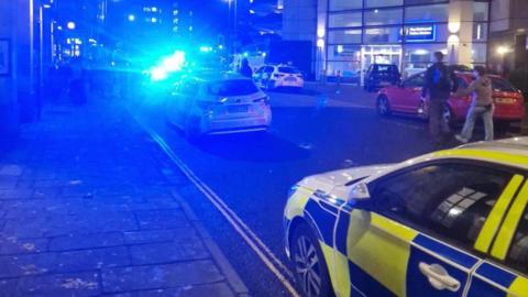 A blurry image of a city centre street with several police cars parked up with their blue lights on. There is a police station in the background and a number of people stood outside.