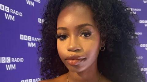 Ms Moponda pictured in a radio studio. There is a banner behind her with the words BBC Radio WM. She is wearing her dark hair is in a long bun. She is wearing a gold necklace with the letter K on and a mustard-colour bandeau top