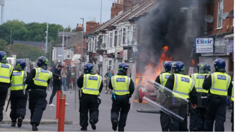 A row of police officers in riot gear, carrying shields walking towards a crowd of men further down the street. A row of shops can be seen on the right and a red car on fire sits in front of them.