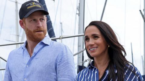 Prince Harry wears a blue shirt looking sideways at Meghan who wears a striped dark blue shirt