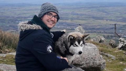 Dean Cooper and his dog Odin on a walk in Cornwall. He is seen at the top of a tor. There is a view of fields behind him.