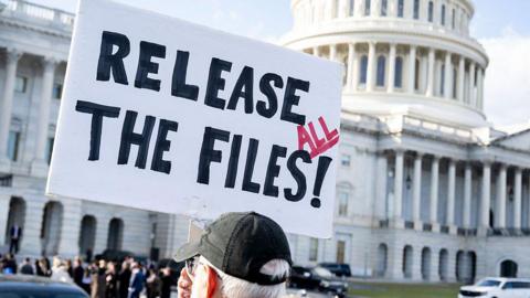 Man holding a sign that says "release all the files" with a black hat on standing outside the US Capitol on 12 November.