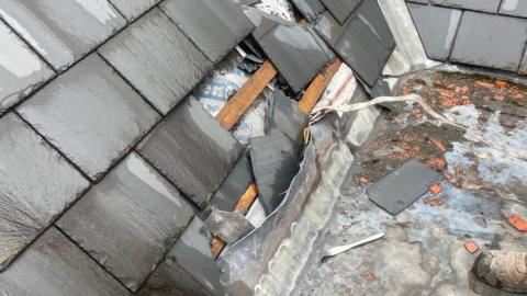 A slate roof has been damaged and the cutlery used to damage it is on the floor. There is slate missing from the roof and it is on the ground. 