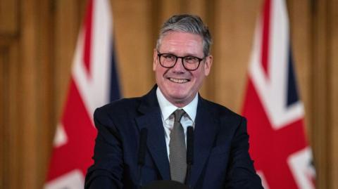Sir Keir Starmer standing in the Number 10 briefing room. He is smiling at the crowd. He wears a dark suit, white shirt, grey tie and black-rimmed glasses. Behind him are two union jack flags.