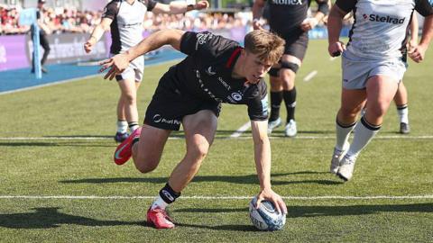 A rugby player wearing black places the ball down over the white line with two opposition players and a teammate watching in the background.